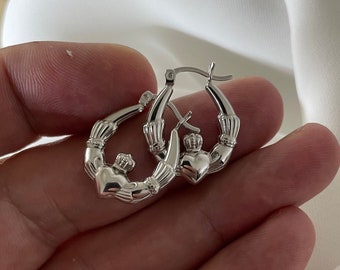 Może przedstawiać: Para srebrnych kolczyków koła z wzorem Claddagh. Każdy kolczyk ma serce z koroną na górze, trzymane przez dwie dłonie.