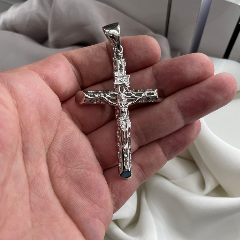 Op de afbeelding: Zilverkleurige hanger met een gedetailleerd kruisbeeld met een figuur van Jezus Christus. De hanger heeft een gestructureerd ontwerp en wordt in een hand gehouden.