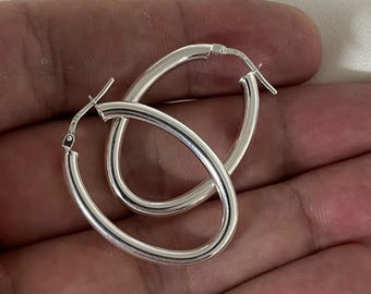 May include: A pair of oval silver hoop earrings. The polished earrings reflect light. The hoops have a classic design, suitable for everyday wear. The earrings are shown against a neutral background. The earrings are made of silver.