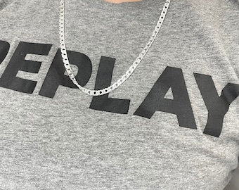 Op de afbeelding: Een zilveren ketting met een curb-link ontwerp. De ketting wordt gedragen door een persoon die een grijs T-shirt draagt met het woord "REPLAY" erop gedrukt.