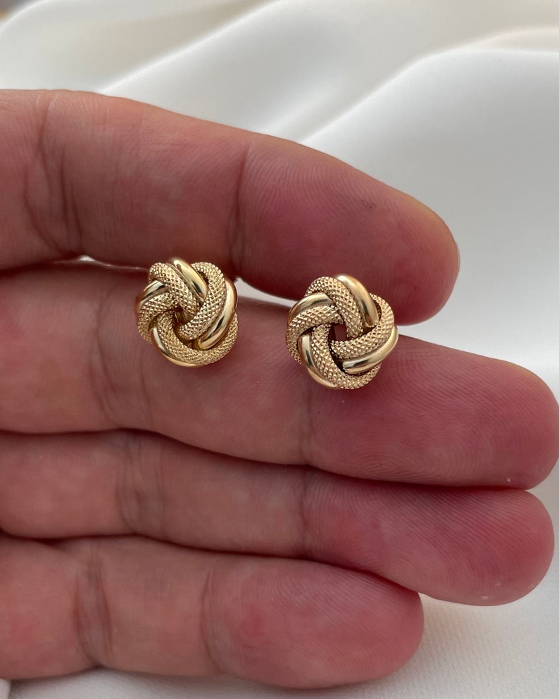 Puede incluir: Un par de pendientes de oro en forma de nudo. Los pendientes son peque&ntilde;os y delicados, y tienen una superficie texturizada.