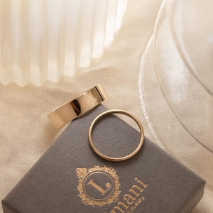 May include: Two gold wedding bands on a grey box with the text "L.mani Jewellery" printed on it.
