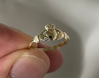 Może przedstawiać: Złoty pierścionek Claddagh z sercem, koroną i dwoma splecionymi dłońmi.