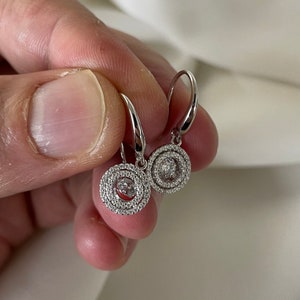 May include: A pair of silver dangle earrings with a round, double-layered design. Each earring features a small, clear gemstone in the center surrounded by smaller, clear gemstones.
