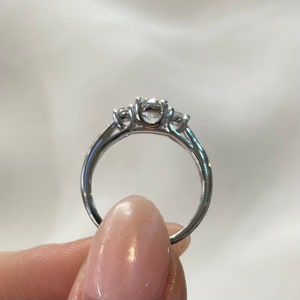 Puede incluir: Un anillo de compromiso de plata de tres piedras con una piedra central m&aacute;s grande y dos piedras m&aacute;s peque&ntilde;as a cada lado.