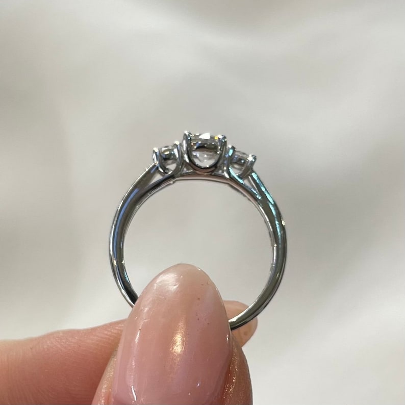 Puede incluir: Un anillo de compromiso de plata de tres piedras con una piedra central m&aacute;s grande y dos piedras m&aacute;s peque&ntilde;as a cada lado.