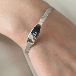 Könnte beinhalten: Ein silberfarbenes Armband mit dem eingravierten Namen "Gia" auf einer ovalen Platte. Das Armband hat ein zartes Blumenmuster. Dieses Schmuckstück ist für das Handgelenk bestimmt.