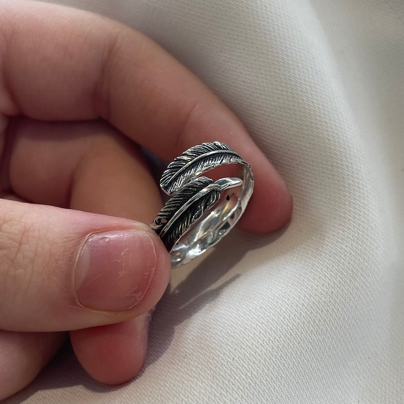 May include: A silver ring with two feather designs. The ring is open and adjustable.