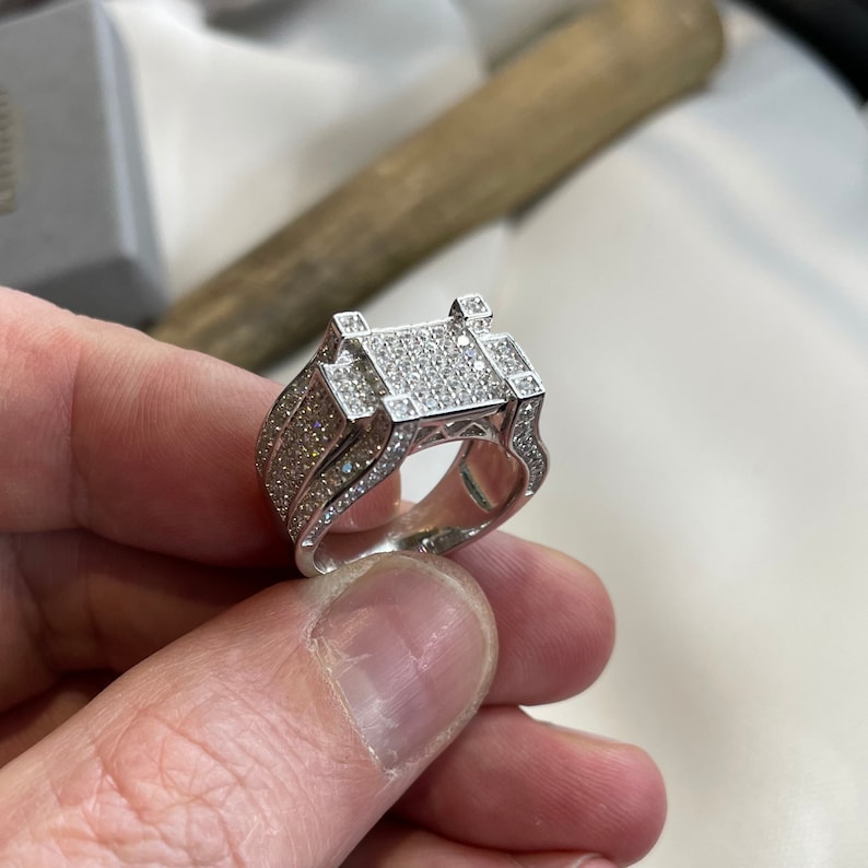 May include: A silver ring with a square-shaped center stone set in a pave setting. The ring is surrounded by smaller diamonds.