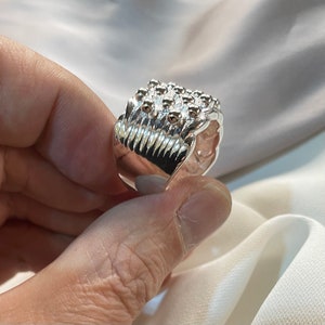 Puede incluir: Un anillo de plata con un diseño geométrico texturizado. El anillo está hecho de múltiples bandas apiladas con una superficie texturizada en relieve.