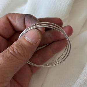 Può includere: Un set di tre braccialetti color argento. I braccialetti sono sottili e rotondi, con una superficie liscia e riflettente. I gioielli sono esposti su uno sfondo bianco morbido.