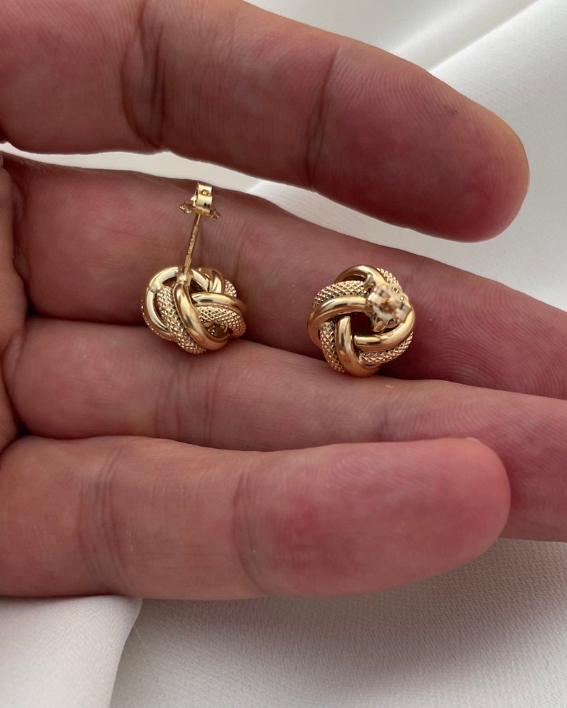 Puede incluir: Un par de pendientes de oro en forma de nudo. Los pendientes son peque&ntilde;os y delicados, y tienen un dise&ntilde;o simple y elegante.