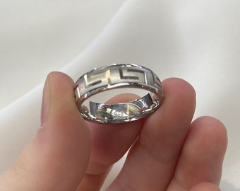 May include: A silver ring with a Greek key design. The ring has a matte finish on the key pattern and a polished band. The ring is held between two fingers.