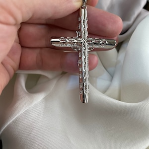 Op de afbeelding: Een zilveren kruis hanger met een gedetailleerd, gestructureerd ontwerp. Het kruis wordt in een hand gehouden, waardoor de ingewikkelde details zichtbaar worden.