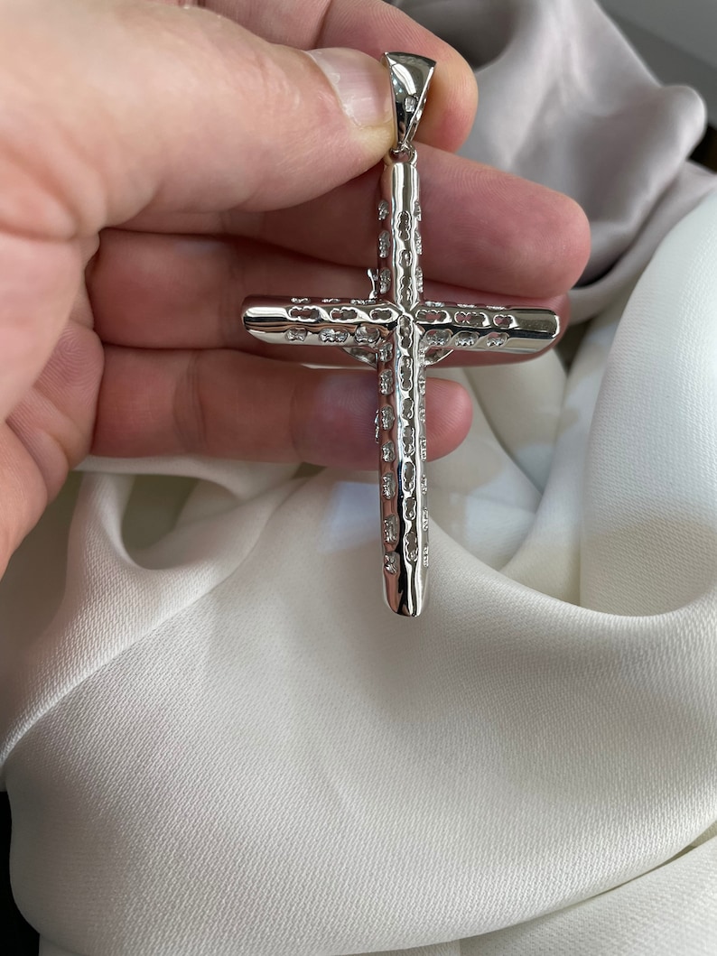 Op de afbeelding: Een zilveren kruis hanger met een gedetailleerd, gestructureerd ontwerp. Het kruis wordt in een hand gehouden, waardoor de ingewikkelde details zichtbaar worden.