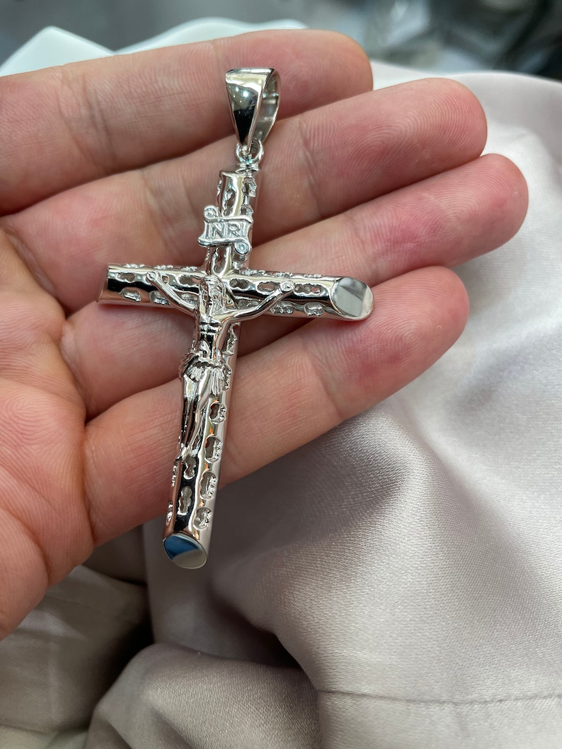 Op de afbeelding: Een zilverkleurige, gedetailleerde, sierlijke kruis hanger met een figuur van Jezus Christus op het kruis. De hanger heeft een lus aan de bovenkant om aan een ketting te bevestigen.