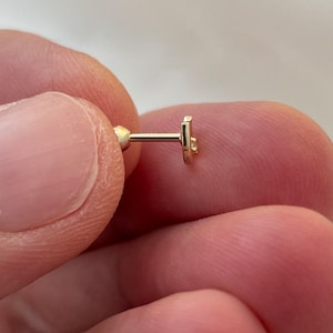 May include: A small, gold-colored earring with a post and a flat, stylized star design. The earring is held between fingers, showcasing its delicate size and intricate details. The earring is designed for ear piercing.