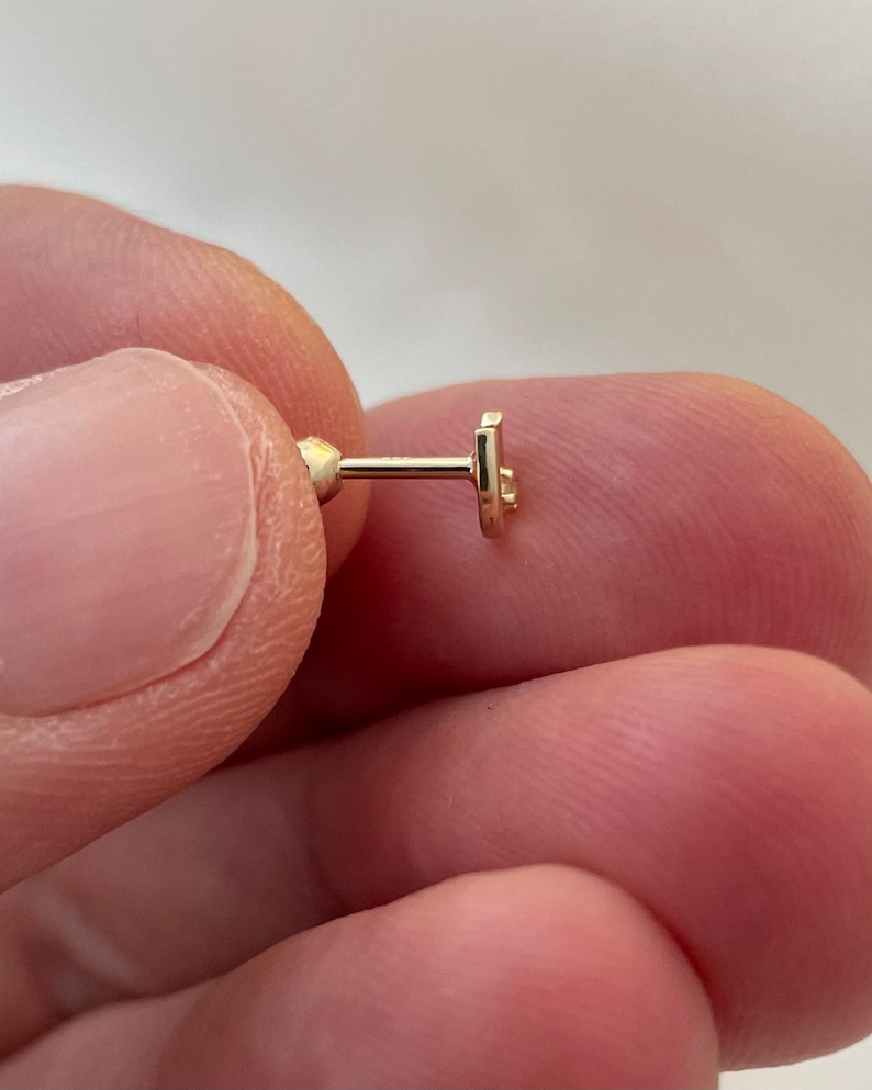 May include: A small, gold-colored earring with a post and a flat, stylized star design. The earring is held between fingers, showcasing its delicate size and intricate details. The earring is designed for ear piercing.