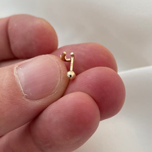 May include: A small, gold-colored earring with a post and a small ball at the bottom. The earring has a curved top and a straight post. The earring is held between two fingers.