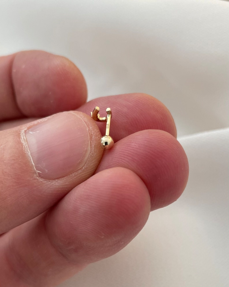 May include: A small, gold-colored earring with a post and a small ball at the bottom. The earring has a curved top and a straight post. The earring is held between two fingers.