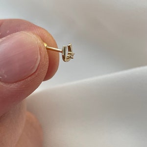 May include: A gold-colored horseshoe-shaped earring with a small, clear gemstone at the center. The earring is held between fingers against a white background, showcasing its delicate design and craftsmanship.