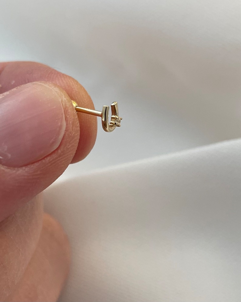 May include: A gold-colored horseshoe-shaped earring with a small, clear gemstone at the center. The earring is held between fingers against a white background, showcasing its delicate design and craftsmanship.