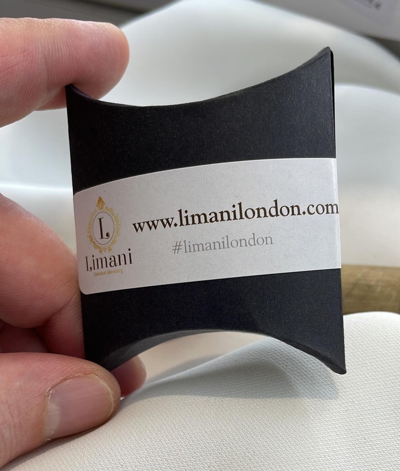 Pu&ograve; includere: Una scatola a cuscino nera con un'etichetta bianca che dice "www.limanilondon.com" e "#limanilondon". L'etichetta presenta anche un logo dorato con la lettera "L" e la parola "Limani".