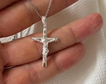 May include: A silver-colored crucifix pendant on a delicate chain. The cross features a detailed depiction of Jesus Christ. The necklace is displayed against a soft, white background.