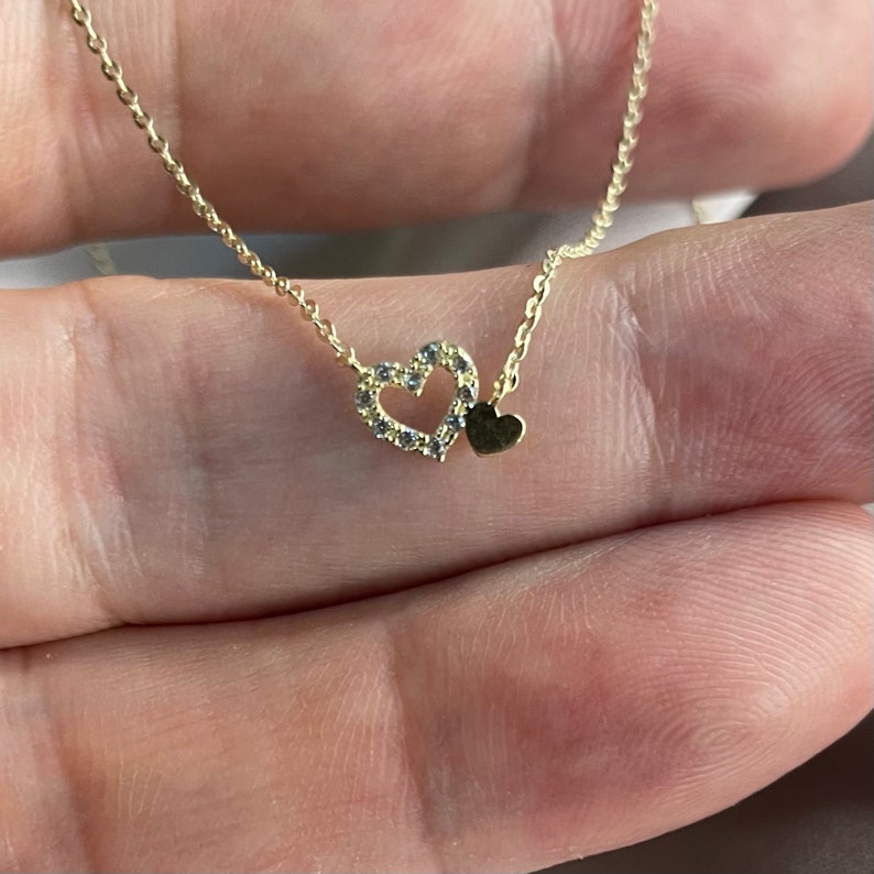 May include: A gold chain necklace with a delicate heart pendant. The heart is outlined with small sparkling stones and has a smaller solid heart charm hanging from it.