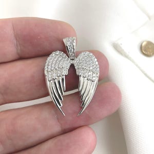 May include: A silver-coloured pendant shaped like angel wings, set with small, sparkling stones. The wings feature detailed feather lines, and the pendant has a small, gem-covered bail for hanging.