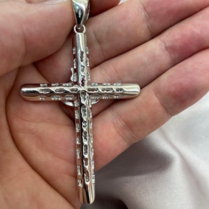 Op de afbeelding: Een zilveren kruis hanger met een gedetailleerd, gestructureerd ontwerp. Het kruis wordt in een hand gehouden, waardoor de grootte en de fijne details worden benadrukt.