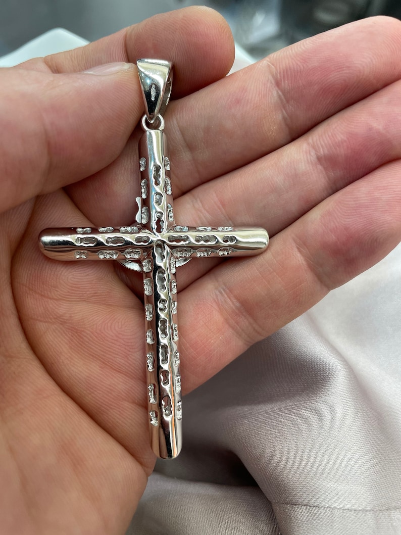 Op de afbeelding: Een zilveren kruis hanger met een gedetailleerd, gestructureerd ontwerp. Het kruis wordt in een hand gehouden, waardoor de grootte en de fijne details worden benadrukt.