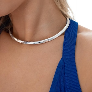 May include: A silver-colored, textured choker necklace. The necklace is worn against a blue top. The choker has a snake chain design, and it sits close to the neck. The background is a light color.