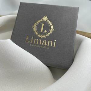 Puede incluir: Una caja de joyer&iacute;a gris con letras doradas y un emblema decorativo. La caja est&aacute; grabada con el nombre de la marca "Limani Classical Jewellery". La caja descansa sobre una tela blanca.