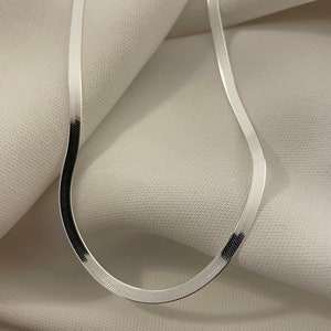 May include: A silver chain necklace with a herringbone pattern. The chain is lying on a white fabric background.