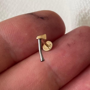 Op de afbeelding: Een kleine gouden en zilveren oorbel in de vorm van een bijl. De bijlkop is goudkleurig, de steel is zilverkleurig en de oorbelpin is goudkleurig. De oorbel wordt tussen twee vingers gehouden.