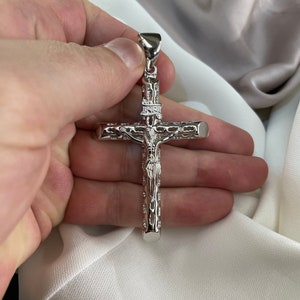 Op de afbeelding: Een zilveren kruis hanger met een gedetailleerd ontwerp van een figuur op het kruis. De hanger wordt in een hand gehouden.