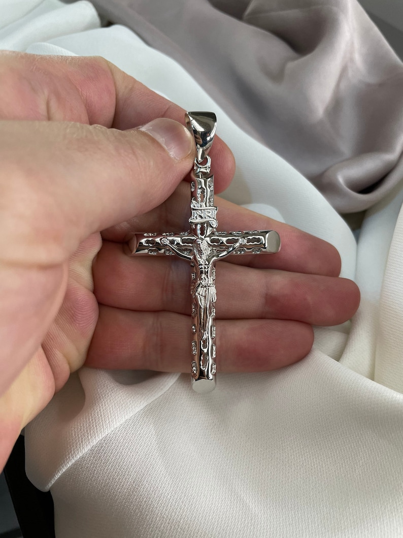 Op de afbeelding: Een zilveren kruis hanger met een gedetailleerd ontwerp van een figuur op het kruis. De hanger wordt in een hand gehouden.