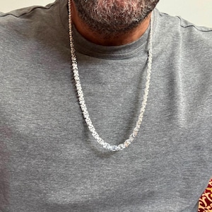 Puede incluir: Un collar de cadena bizantina en tono plateado con un patrón complejo de eslabones entrelazados. La cadena metálica descansa sobre una camiseta gris lisa, mostrando una superficie pulida y reflectante con un diseño cilíndrico constante.