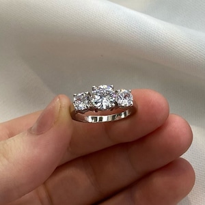 Può includere: Un anello d'argento con tre pietre rotonde trasparenti. L'anello è contrassegnato "925" sulla fascia interna.