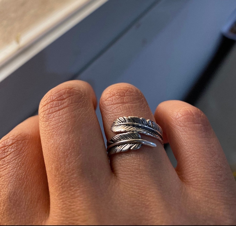 May include: A silver ring with a feather design. The ring is worn on a person's finger.