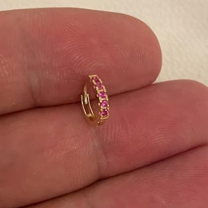 May include: A small, gold-coloured hoop earring with a row of pink gemstones. The earring is held in a person's hand, showcasing its delicate design and vibrant colour. The gemstones are evenly spaced along the front of the hoop.