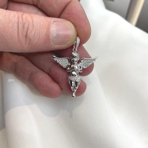 Puede incluir: Un colgante plateado con forma de querub&iacute;n con alas extendidas. El querub&iacute;n est&aacute; detallado con peque&ntilde;as piedras brillantes. El colgante est&aacute; sostenido por una mano sobre un fondo blanco.