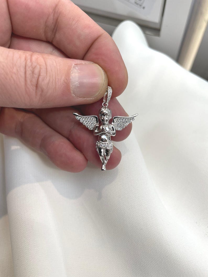 Puede incluir: Un colgante plateado con forma de querub&iacute;n con alas extendidas. El querub&iacute;n est&aacute; detallado con peque&ntilde;as piedras brillantes. El colgante est&aacute; sostenido por una mano sobre un fondo blanco.