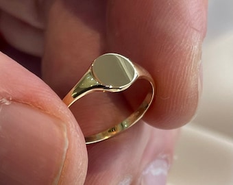 May include: A gold signet ring with a smooth, oval-shaped top. The ring is worn on a finger.
