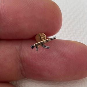 May include: A small, gold-colored stud earring featuring a detailed miniature rifle design. The rifle is black and silver, with intricate detailing. The earring post is gold. The overall design is a miniature representation of a firearm.