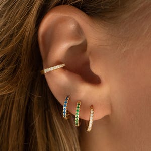 May include: Gold-toned ear cuff and three hoop earrings. The ear cuff is adorned with small, clear stones. The hoop earrings feature blue, green, and clear stones. The jewelry is worn on an ear, showcasing the pieces' design and color.