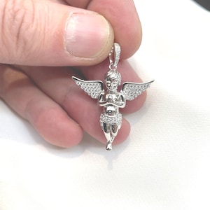 Puede incluir: Un colgante plateado con forma de querub&iacute;n con alas extendidas. El querub&iacute;n est&aacute; adornado con peque&ntilde;as piedras brillantes. El colgante es sostenido por una mano sobre un fondo blanco. Es una pieza de joyer&iacute;a religiosa o simb&oacute;lica.