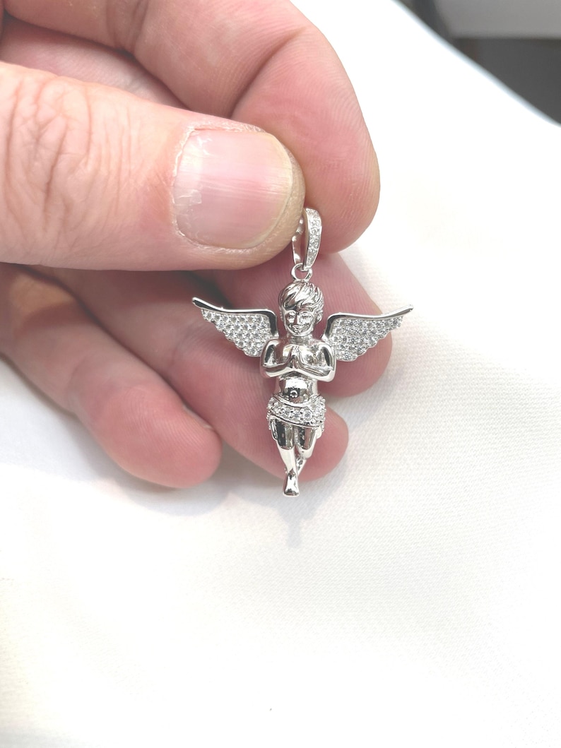 Puede incluir: Un colgante plateado con forma de querub&iacute;n con alas extendidas. El querub&iacute;n est&aacute; adornado con peque&ntilde;as piedras brillantes. El colgante es sostenido por una mano sobre un fondo blanco. Es una pieza de joyer&iacute;a religiosa o simb&oacute;lica.