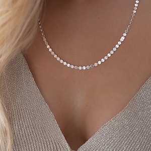 Puede incluir: Un delicado collar de plata con una serie de pequeños discos redondos y planos. El collar se lleva alrededor del cuello. El fondo es de color neutro, resaltando la joya.